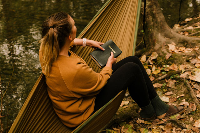 A woman is holding a bible on a hammock in the woods.